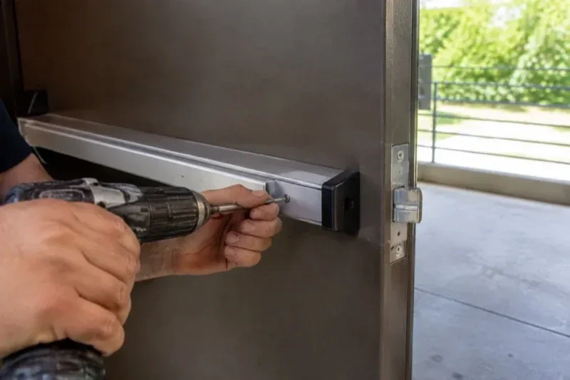 Hands using a power driver at the end of a horizontal push bar on an open metal door. — Panic & Exit Hardware, Ajax
