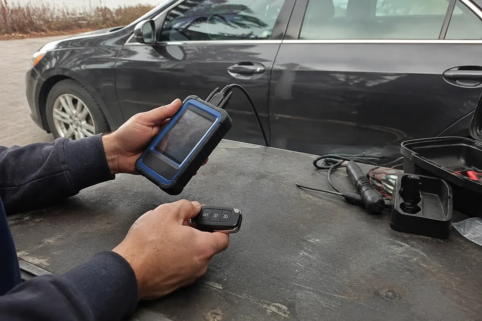 Hands holding a blue-edged electronic device and car-key fob at a worktable beside a parked car. — Transponder Key Programming, Ajax